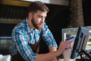 Handsome concentrated bearded waiter swiping credit card through the computer terminal in cafe