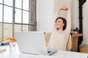 Happy relaxed young woman sitting in her kitchen with a laptop in front of her stretching her arms above her head and looking out of the window with a smile-2