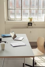 Laptop, documents and a paper coffee cup on a white desk in the interior of a modern bright office with large window, radiator and a wooden chair on wheels-1